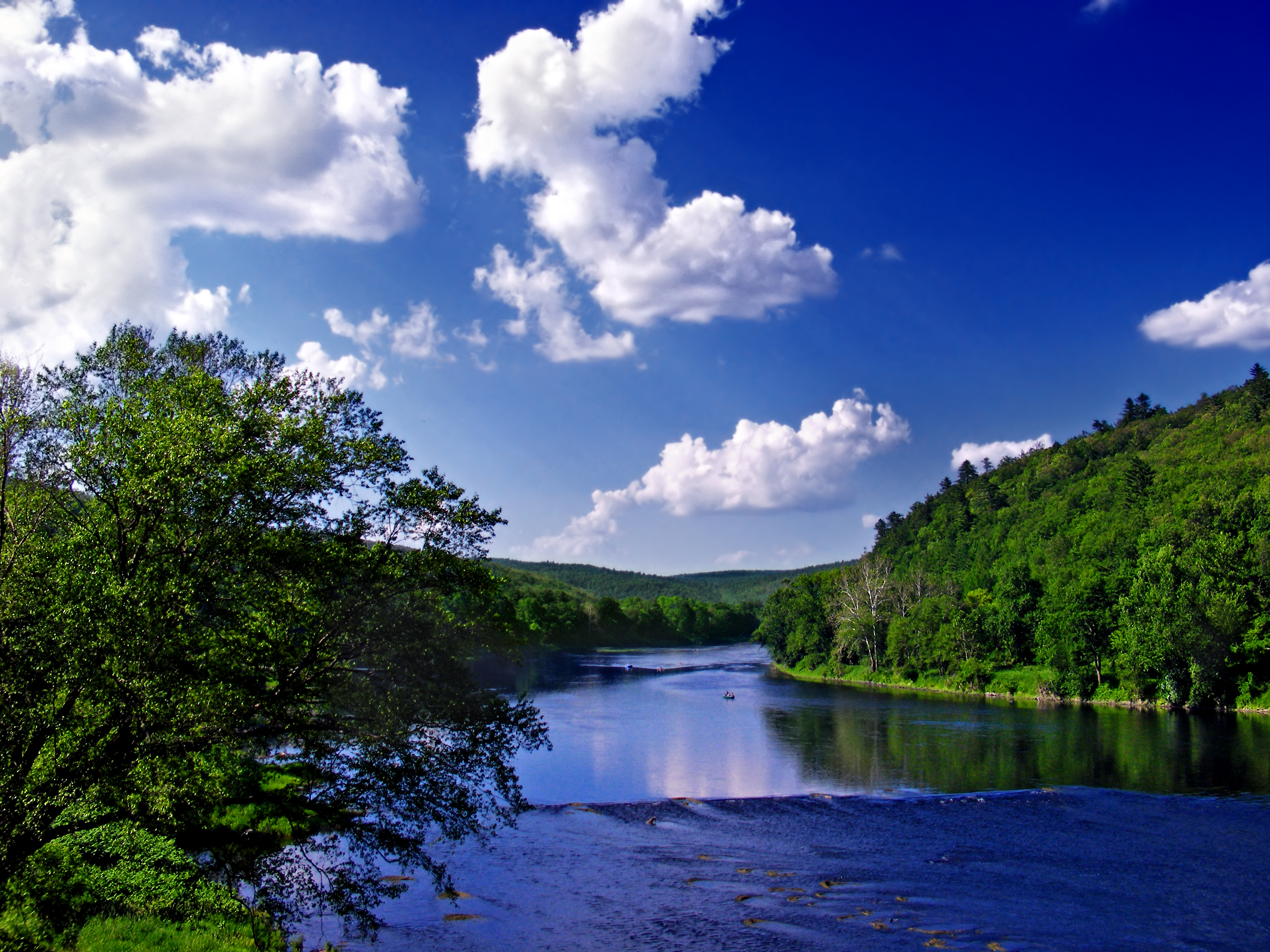 A river runs through rolling, forested terrain under blue sky with puffy clouds.
